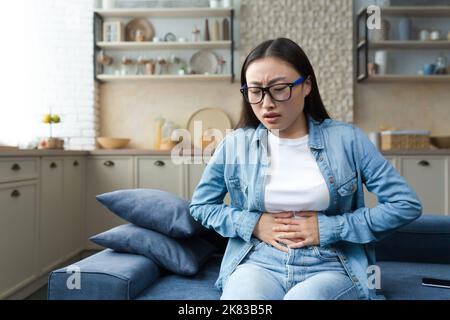Stomach disorder. A young beautiful Asian woman holds her stomach with her hands, feels severe pain. Sitting at home on the sofa. Stock Photo