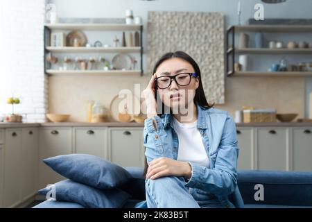 Depression. Tired and pensive young Asian woman sitting on sofa at home, holding hand to head, looking focused. Stock Photo