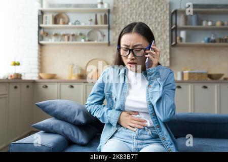 Scared and anxious young pregnant Asian woman sitting on the sofa at home and holding her stomach, feeling severe pain, cramp in her stomach. He calls an ambulance, calls the hospital. Stock Photo