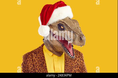 Man in funny dinosaur mask, red Christmas hat and leopard jacket isolated on yellow background Stock Photo