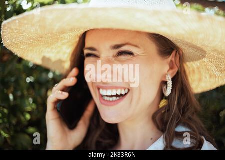 happy stylish middle aged housewife in white shirt with hat using a smartphone outdoors near green wall. Stock Photo