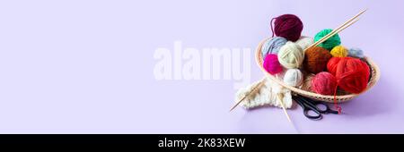 Red, yellow, white, purple, brown, green balls of thread in a beige basket with knitting needles made of bamboo and scissors on a lilac background, ba Stock Photo