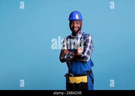 Optimistic african american builder using tool belt and cordless power drill. Contractor proud of skills looking confidently at camera in studio isolated on blue background. Stock Photo