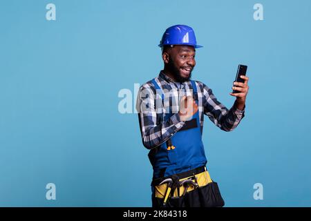 African american builder happy for news of renovation job received by cell phone message. Contractor in uniform excited using mobile device in studio shot against blue background. Stock Photo