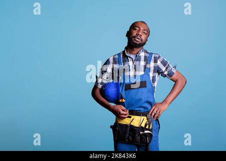African american construction worker looking seriously at camera in studio shot against blue background. Construction worker at the end of the workday holding hard hat under arm. Stock Photo