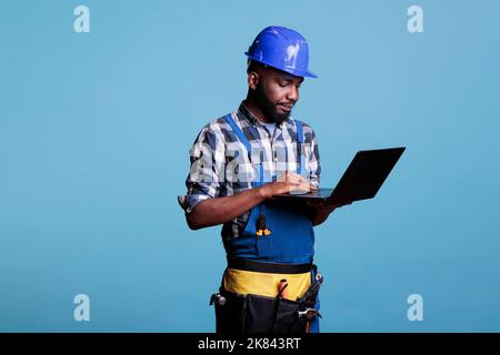 Construction worker in work uniform and hard hat using modern laptop computer. Professional male construction site supervisor wearing a tool belt while using a computer on a blue background. Stock Photo
