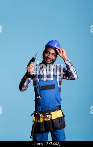 Happy african american construction worker adjusting hard hat holding electric cordless drill. Optimistic handyman wearing tool belt in studio shot isolated on blue background. Stock Photo