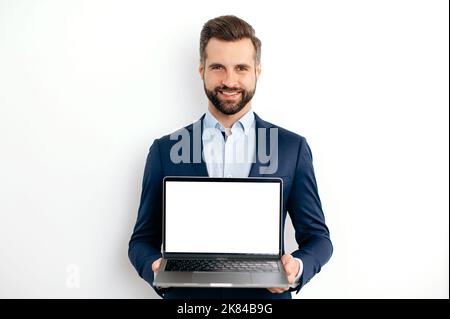 Advertisement concept. Positive caucasian bearded business man, in suit, programmer, IT specialist, seo, holds open laptop with empty mock-up screen in hand, stand on isolated white background, smiles Stock Photo
