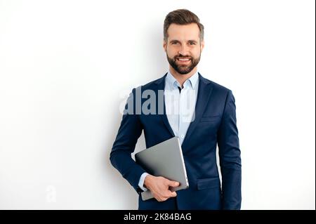 Portrait of a positive successful caucasian bearded business man in a suit, seo, consultant, broker, standing on isolated white background, holding laptop, looking at camera, smiling friendly Stock Photo