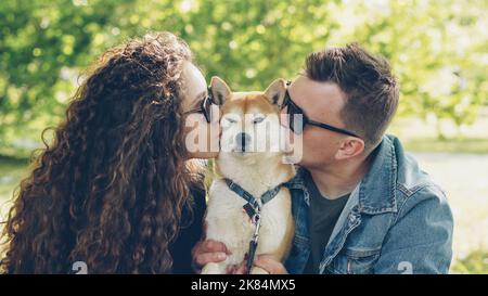 Loving owners of beautiful dog shiba inu are kissing the animal and petting it on the head while resting in the park together. Love, pets, people and healthy lifestyle concept. Stock Photo