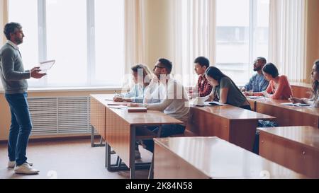 Male professor in casual clothes is talking to group of students sitting at tables in classroom and making notes. Large lecture hall with desks, chairs and large windows is visible. Stock Photo