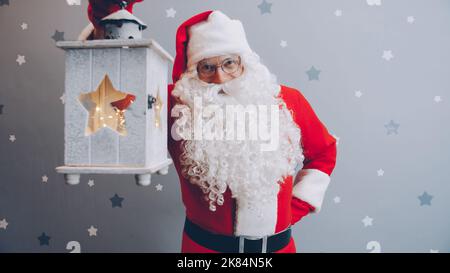 Portrait of man in Santa suit holding lantern and smiling looking at camera on starry grey background. Holidays and festivals concept. Stock Photo