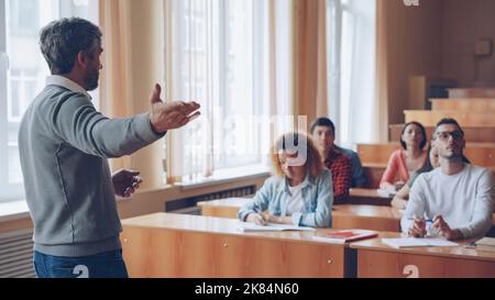 Bearded mature man professor is reading lecture talking and gesturing while students are listening and writing sitting at tables in spacious university classroom. Stock Photo
