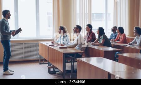 Male professor in casual clothes is talking to group of students sitting at tables in classroom and making notes. Large lecture hall with desks, chairs and large windows is visible. Stock Photo