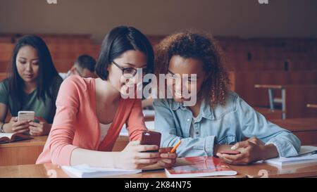 Pretty girls students students are using smartphone, watching screen, talking and laughing sitting at desks at university. Social media, internet, millennials and education concept. Stock Photo