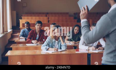Group of students is listening to teacher and writing sitting at tables in classroom while male professor is talking and gesturing. Higher education and people concept. Stock Photo