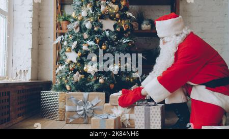 Santa Claus in traditional outfit is putting gifts under Christmas tree on holiday at home, room is beautifully decorated. Occasions and presents concept. Stock Photo