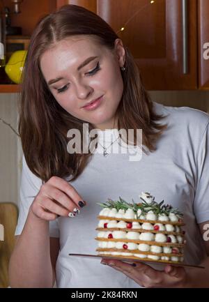 A beautiful happy girl in a white T-shirt, stands at home, holds a festive New Year's cake on a plate Stock Photo