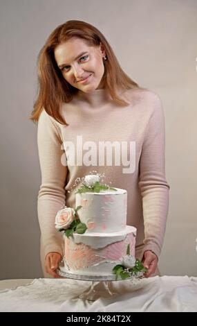 A beautiful girl confectioner shows a cooked two-tiered wedding cake decorated with fresh flowers. Stock Photo
