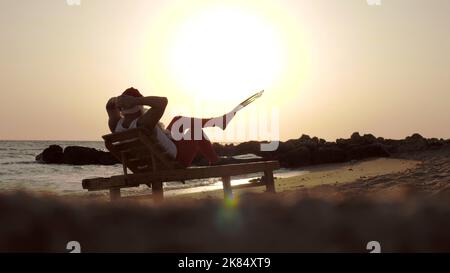 Santa Claus summer vacation. Santa Claus sunbathes. back view. Funny Santa, in flippers, is sitting on lounger, on beach by the sea and enjoying the sunset or sunrise, relaxing. at the seashore. High quality photo Stock Photo