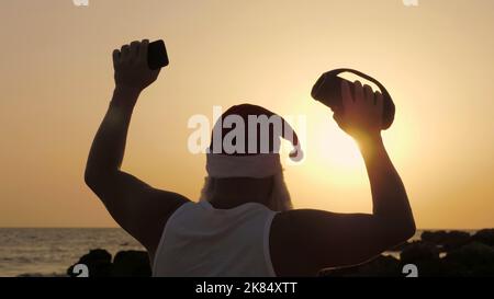 Santa Claus dances. Funny Santa Claus dancing with a wireless musical bluetooth speaker in hand, against backdrop of sunset or sunrise. back view. relax on beach by the sea. High quality photo Stock Photo