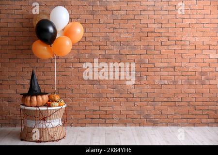 Table with Halloween pumpkins, witch hat and balloons near brick wall Stock Photo