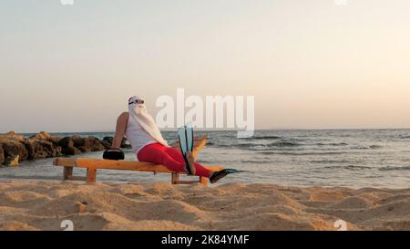 funny santa claus sunbathes. santa, in sunglasses and flippers, is sitting on lounger, on beach by the sea and listening to music, relaxing. santa claus summer vacation, at the seashore. High quality photo Stock Photo