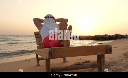 funny santa claus sunbathes. santa, in sunglasses, lies on wooden lounger on beach by the sea, relaxing. santa claus is on summer vacation, at the seashore. High quality photo Stock Photo