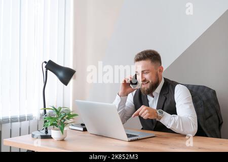 Happy male employee in suit with beard sitting at workplace using laptop smiling on smartphone, positively cheerful man in court with computer chatting on mobile phone with friend in office Stock Photo