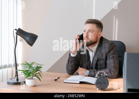 Pensive bearded young man calling by smartphones and writing down notes in notebook while sitting at a table in the office. Serious hipster guy taking notes while having a video call Stock Photo