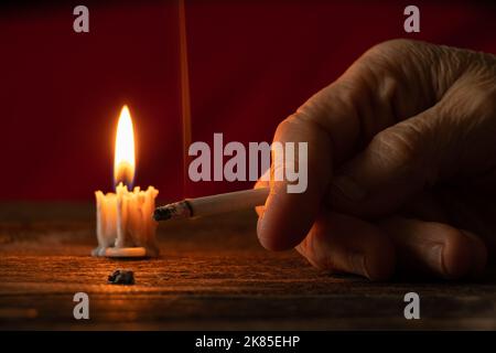 old woman holding a cigarette by candlelight in the dark at home at the table, smoking at home in the dark, nicotine and bad habit, cigarettes and can Stock Photo