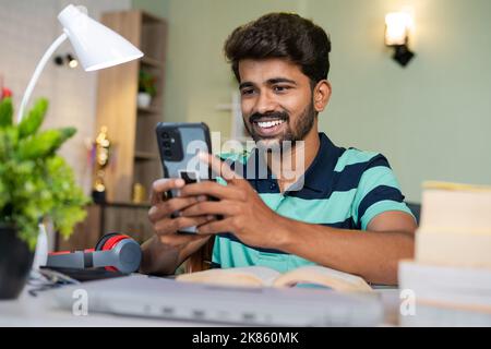 happy smiling college Student using mobile phone while reading at study desk - concept of relaxation, taking break and social media addiction Stock Photo