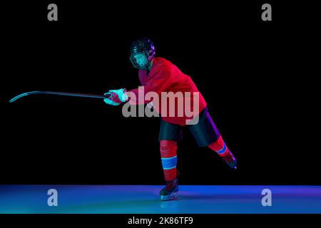 Junior ice hockey player in sports uniform and protective equipment in action over dark background in neon light. Sport, power, challenges Stock Photo