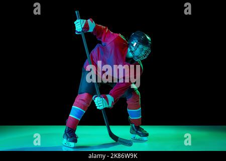 Junior ice hockey player in sports uniform and protective equipment in action over dark background in neon light. Sport, power, challenges Stock Photo