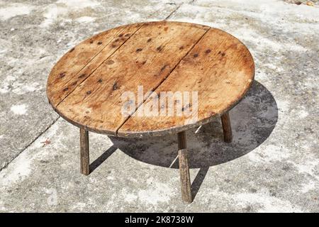 Handmade wooden table with three legs, in a concrete paved courtyard. Stock Photo