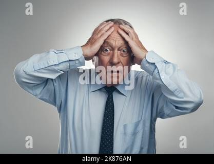 What on earth are you doing. a senior businessman standing against a grey studio background with his head in his hands and looking stressed. Stock Photo