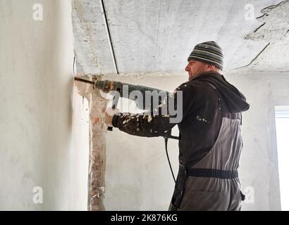 Male worker drilling wall with construction electric drill. Man using professional drilling tool while breaking wall in apartment during renovation. Concept of home renovation and demolition work. Stock Photo