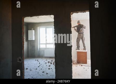 Man builder standing on wooden table and drilling wall between two rooms with doorways. Male worker using electric hammer drill while breaking the wall in apartment during renovation. Stock Photo
