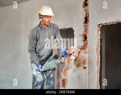 Worker in protective helmet with perforator completing deconstruction of rest of brick wall inside room. Builder carring out repair work on dismantling of walls and partitions. Stock Photo