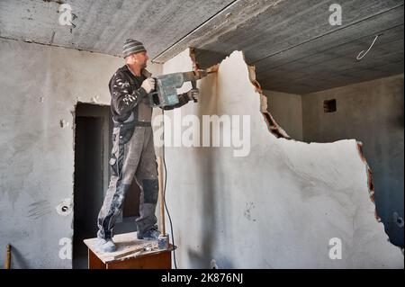 Full length of man in workwear using hammer drill while breaking wall in apartment during renovation. Male worker standing in wooden table and drilling wall with special tool. Stock Photo