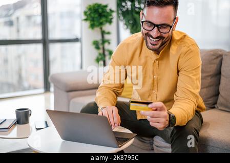 Delighted handsome adult man with eyeglasses, smiling and reading the number on his credit card, at home. Stock Photo
