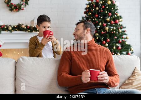 cheerful man sitting on couch and looking at son drinking warm cocoa near decorated christmas tree Stock Photo