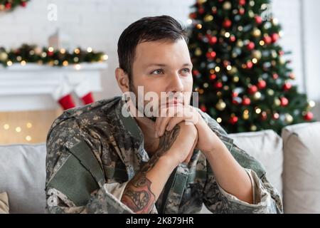 thoughtful man in camouflage sitting with hands near face and looking away near blurred christmas tree Stock Photo