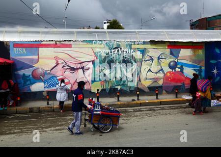 Street seller walking past mural showing Pachamama (Mother Earth) as a sad old woman flanked by Donald Trump and Xi Jinping, La Ceja, El Alto, Bolivia Stock Photo