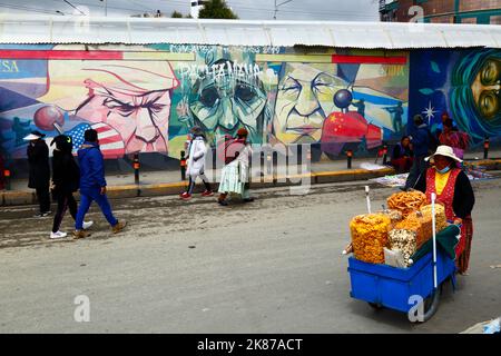 Street seller walking past mural showing Pachamama (Mother Earth) as a sad old woman flanked by Donald Trump and Xi Jinping, La Ceja, El Alto, Bolivia Stock Photo