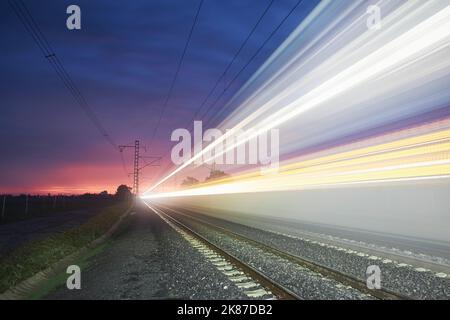 Modern railway at beautiful sunrise. Light trail of high speed train on railroad track. Moving modern intercity passenger train. Stock Photo