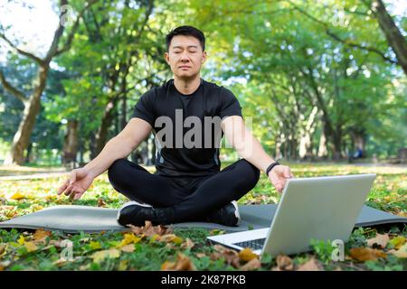 Asian meditating exercise in lotus pose, man resting after fitness training using laptop for online meditation training. Stock Photo
