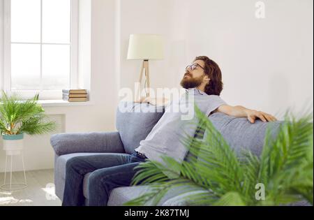 Tired relaxed Caucasian man closed eyes and spread arms to sides sits on sofa after hard day at work Stock Photo