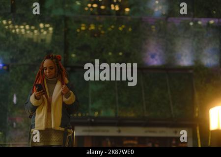 Female hipster with smartphone at night in city. Low angle of serious young woman with dreadlocks browsing mobile phone while standing in city at nigh Stock Photo