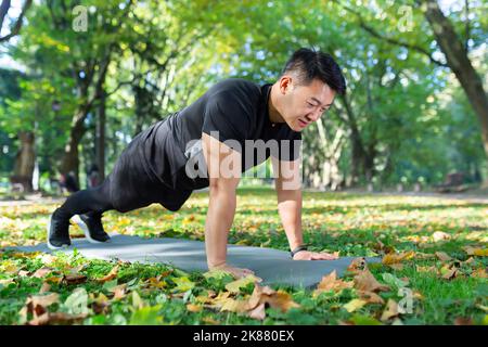 Asian athlete doing push-ups in park and doing plank on sports mat, man happy with workout on sunny day. Stock Photo